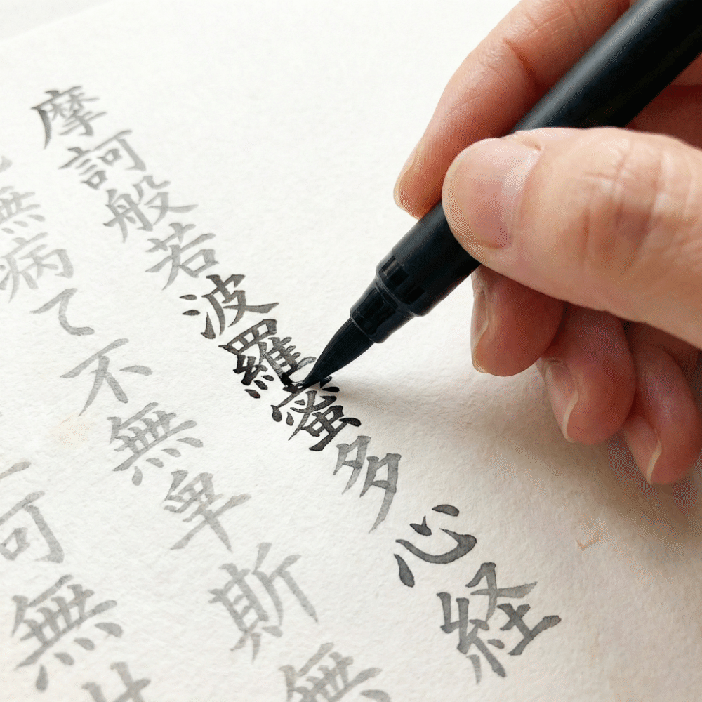 Close-up of hands tracing printed Heart Sutra text with a brush pen during shakyo practice